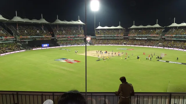 A lit cricket stadium during a night match, viewed from the stands.