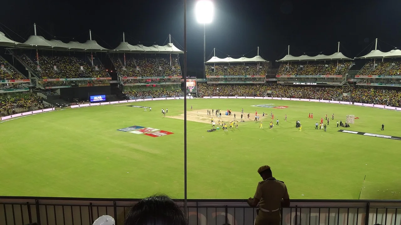 A lit cricket stadium during a night match, viewed from the stands.